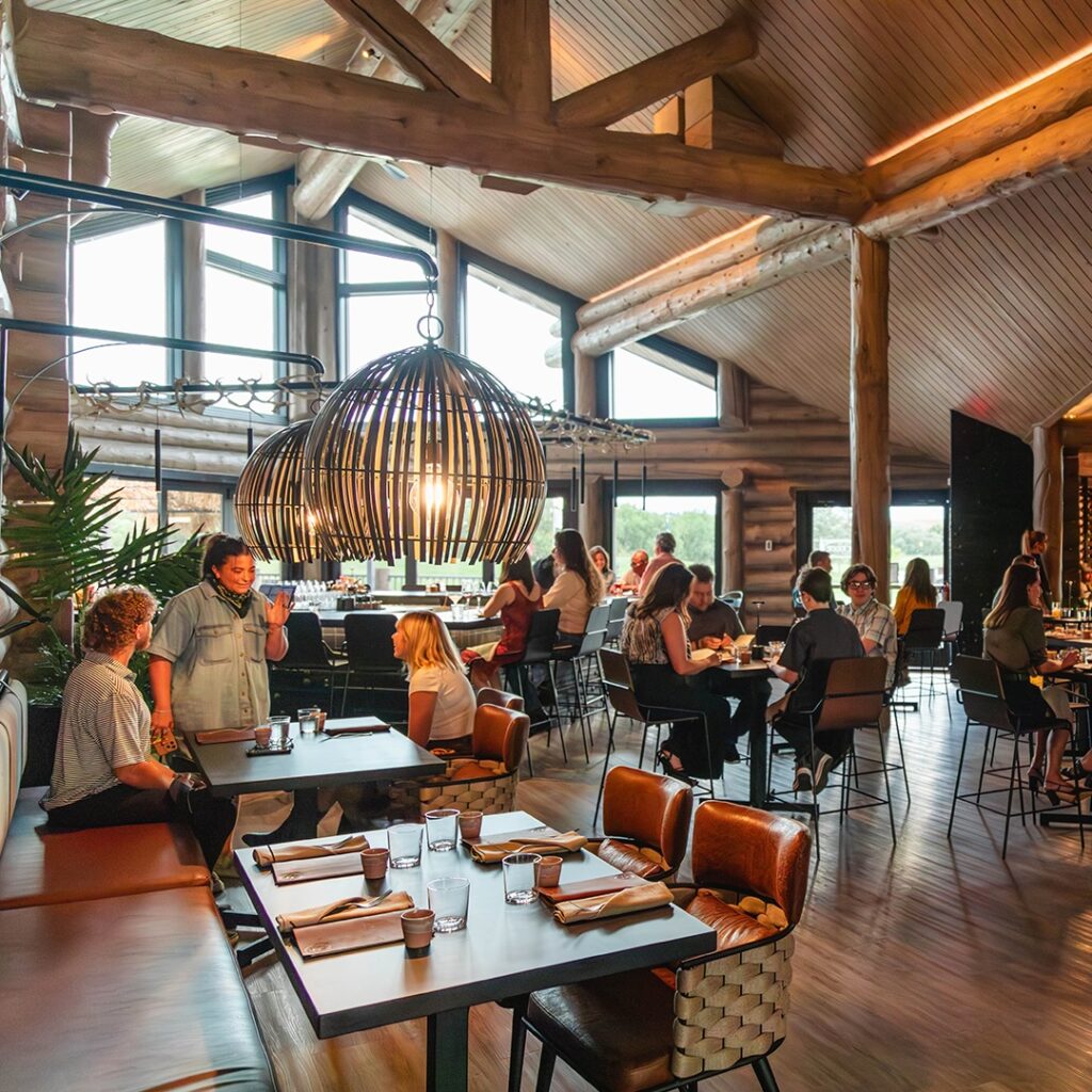 A warm and inviting fine dining restaurant interior with rustic wooden beams, large windows, and modern lighting fixtures. Guests are seated at various tables, enjoying unique culinary experiences in a lively atmosphere.